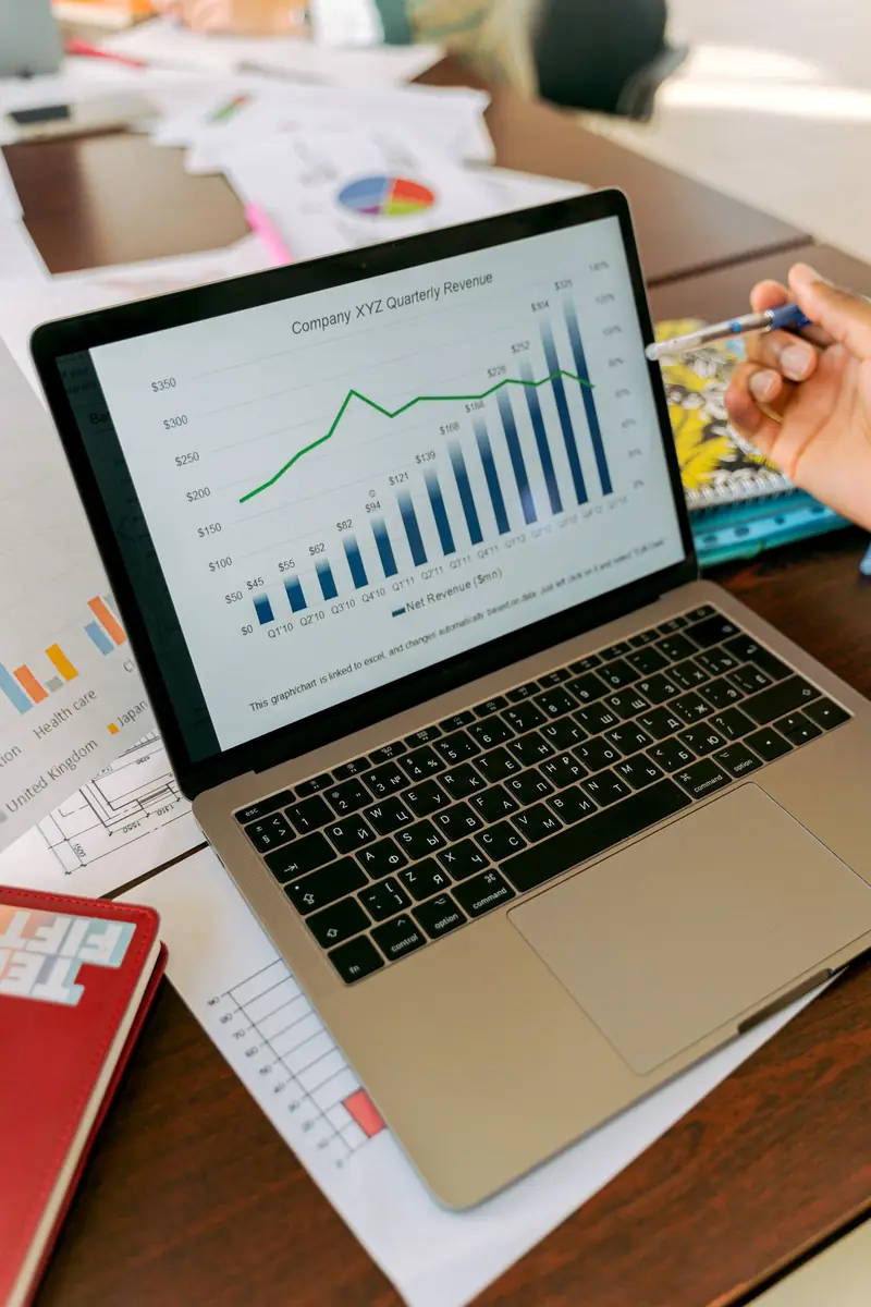 Laptop on a desk showing a quarterly revenue bar chart, with printed reports nearby